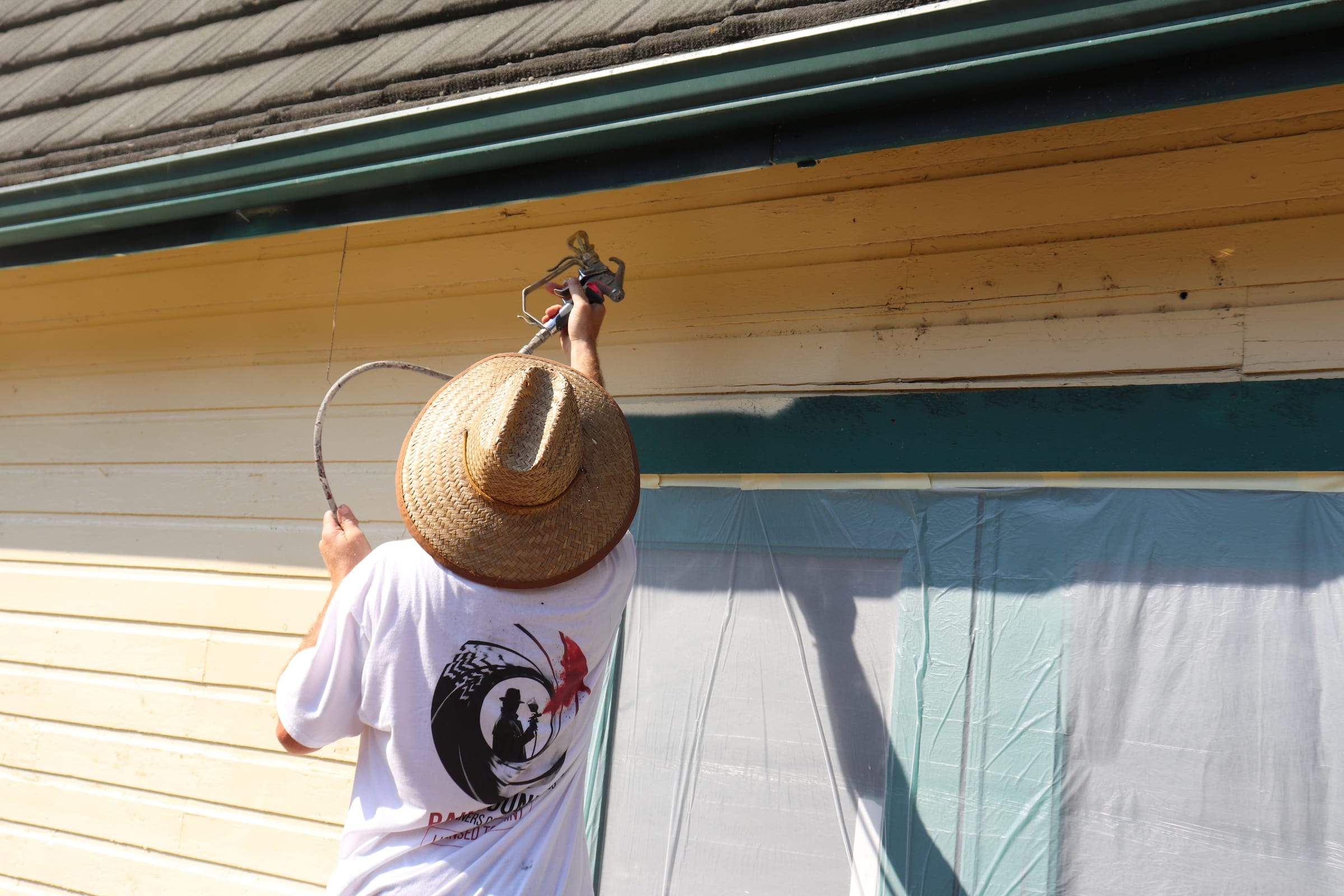 Painter spraying a deep teal fascia band beneath the roof eave of a cream building